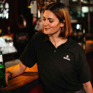 weibliches Model trägt schwarzes Poloshirt mit Becks Logo und grünem Knopf