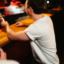Load image into Gallery viewer, männliches Model trägt weißes T-Shirt mit gekrempelten Ärmeln und Becks Schlüssel darauf in Bar Atmophäre