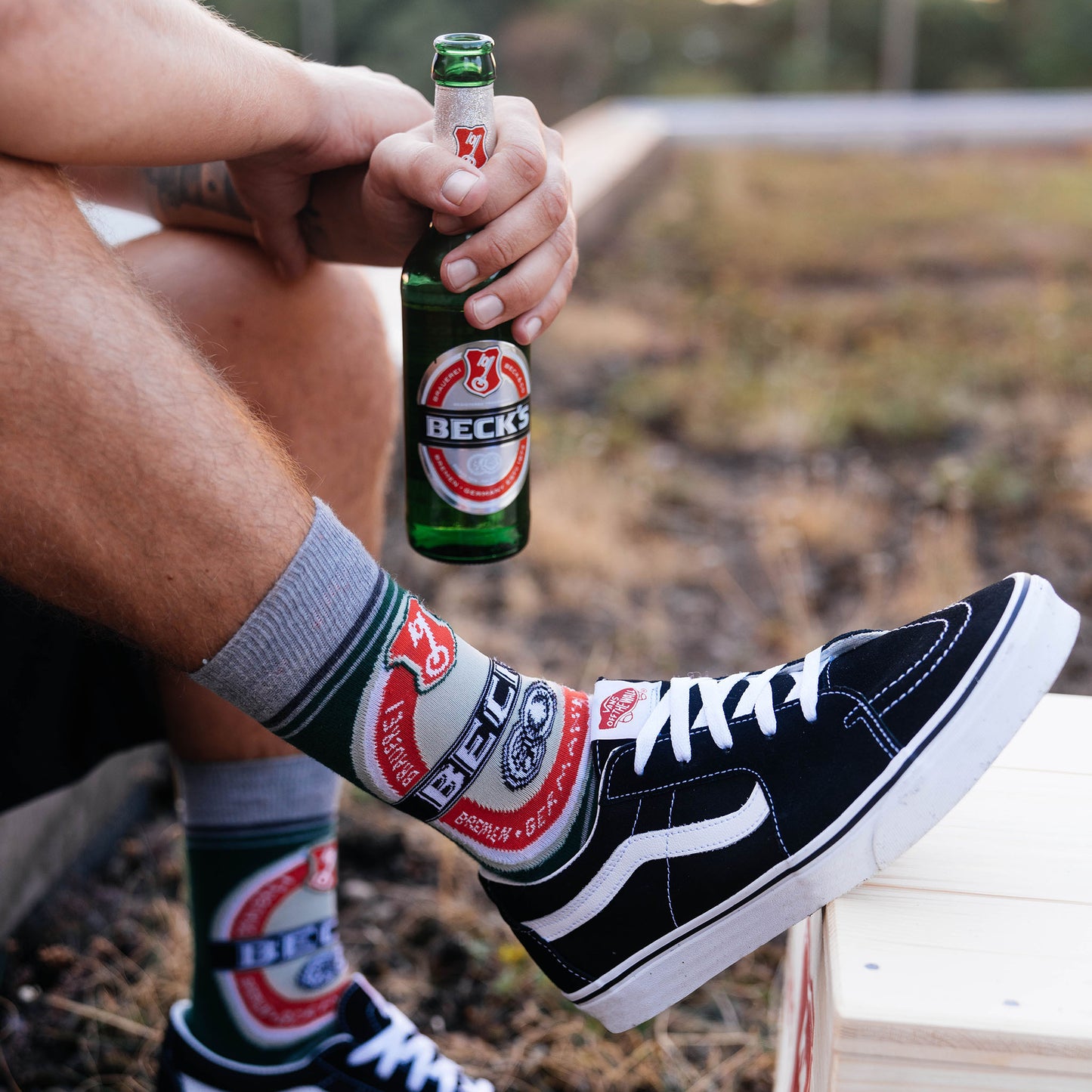 Model mit Becksflasche in der Hand und Füße in schwarzen Vans und hohen Becks Retro Socken auf der Wiese